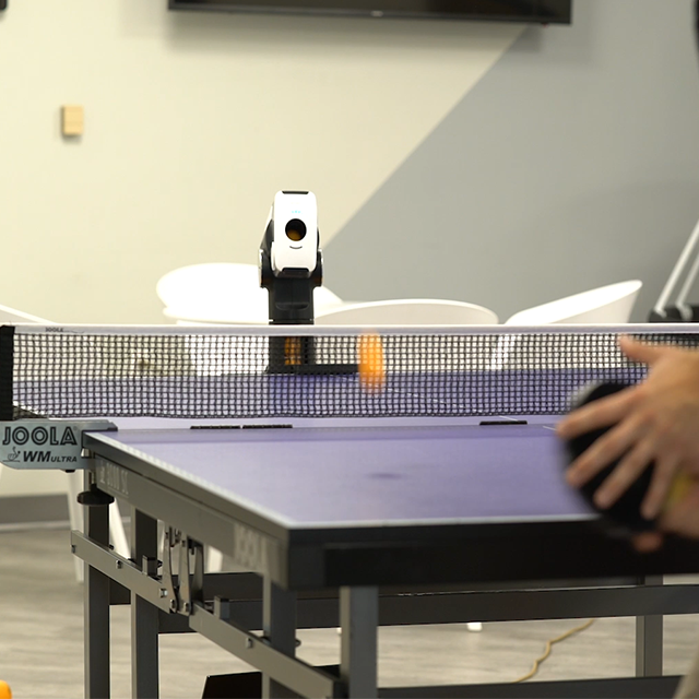 Table tennis robot shooting an orange ball toward a player on a JOOLA table, ideal for precision training drills.